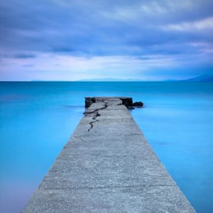 Broken concrete pier or jetty and rocks on a blue sea. Hills on background. Long exposure photography.